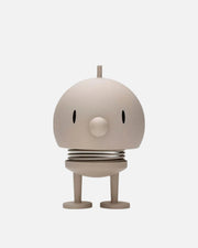 A minimalist, stylized robot figurine in a warm beige or cream color, featuring a rounded dome head with small black oval eyes, a circular nose, a spring-like ribbed waist, and two short legs on a plain white background.