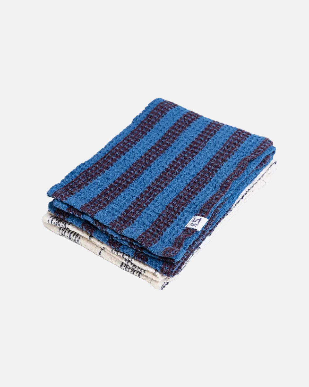 A folded waffle-weave textile, likely a blanket or towel, featuring alternating stripes of medium blue and dark navy, with a patterned cream and blue reverse side visible on the underside. A small white tag is visible on the side.