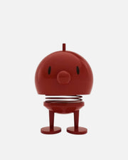 A red spring toy figure with a round head, simple black eyes, a protruding top stem, and two short legs on a plain white background.